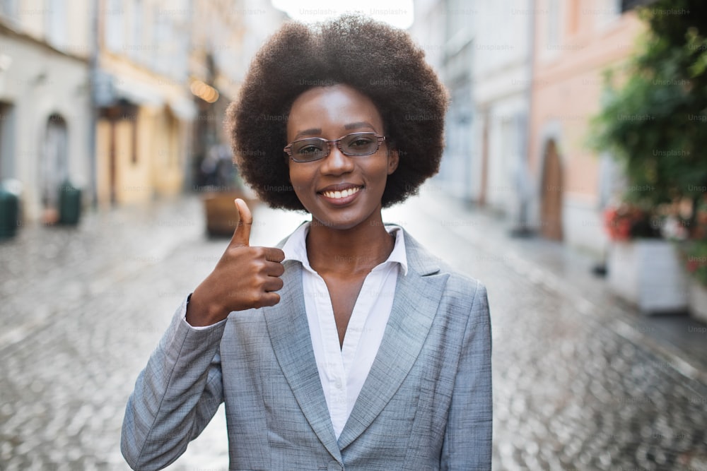 眼鏡をかけ、屋外で親指を立てるフォーマルな服を着た笑顔のアフリカ人女性。自信と成功したキャリアのコンセプト。