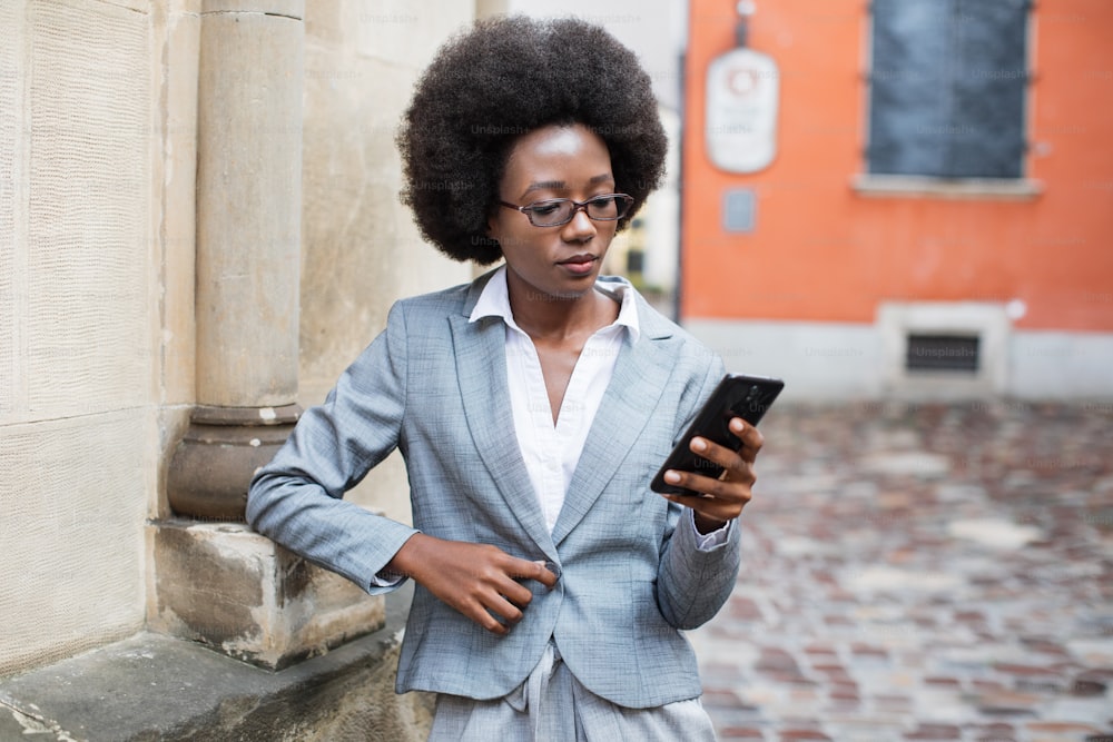 Attractive business woman with dark skin standing on city street with clipboard and modern smartphone in hands. Busy lady in suit and eyeglasses solving working issues online.