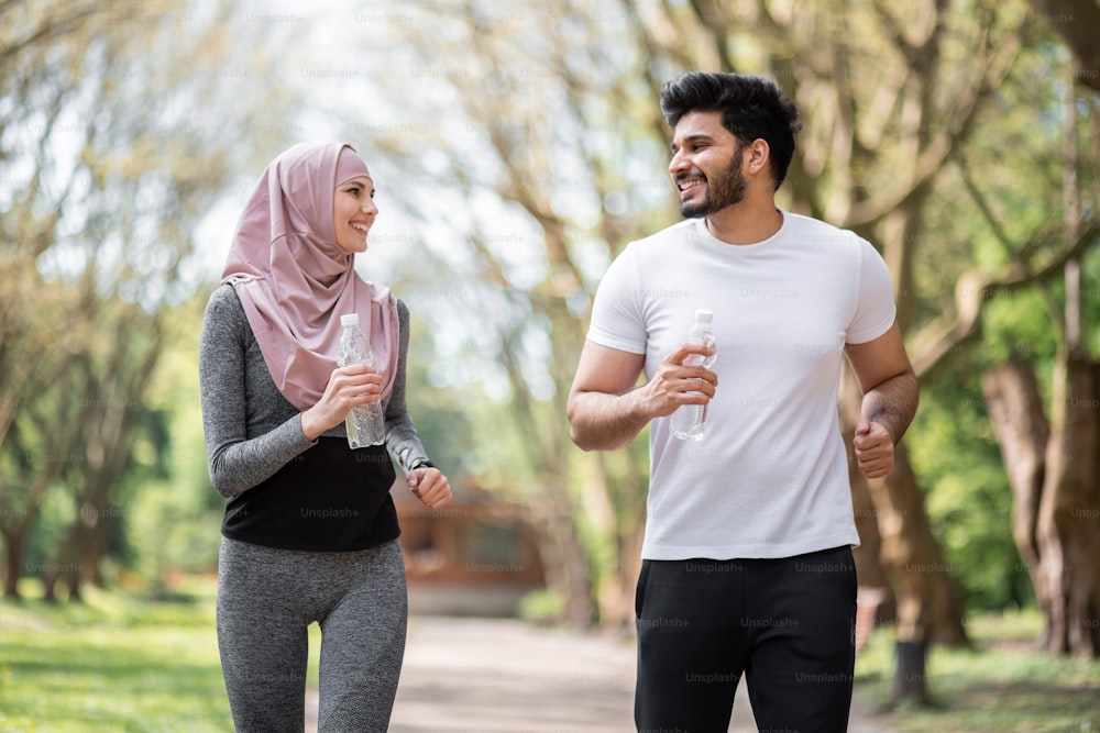 Coppia araba felice in abbigliamento sportivo che corre insieme al parco estivo con una bottiglia d'acqua in mano. Giovane uomo e donna che trascorrono il tempo libero attivamente all'aria aperta.