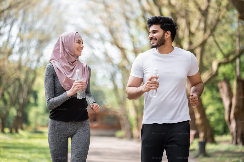 Happy arabian couple in activewear running together at summer park with bottle of water in hands. Young man and woman spending free time actively on fresh air.