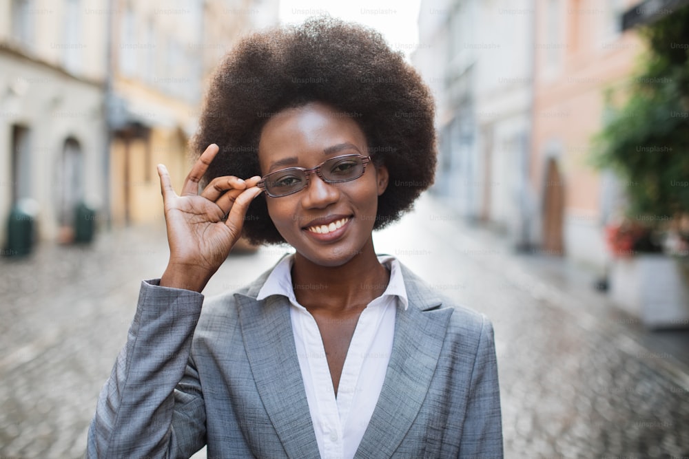 Portrait of positive afro american lady in stylish business suit touching with hand her eyeglasses while standing outdoors. Successful woman smiling and looking at camera on street.