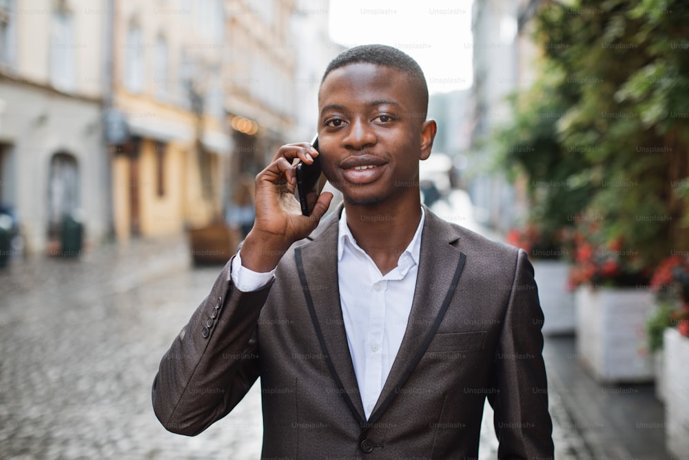 Confident businessman in stylish suit having working conversation on mobile while standing on street. Handsome african man using smartphone for talk outdoors.