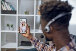 Vue de l’épaule d’un homme afro-américain dans un casque ayant une leçon en ligne sur smartphone avec une enseignante caucasienne. Concept de personnes, de technologie et d’enseignement à distance.