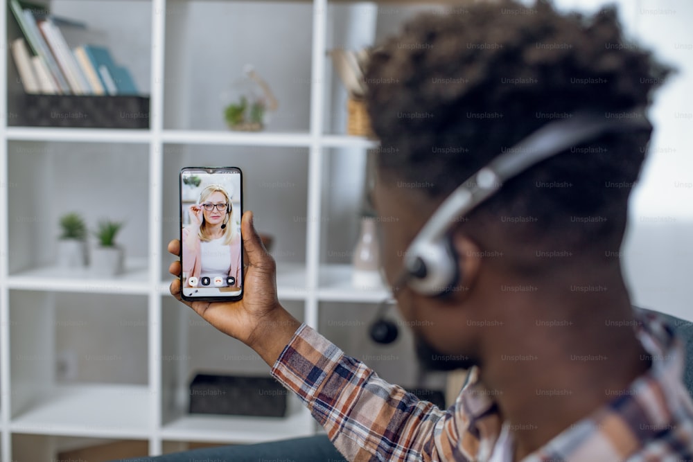 View from shoulder of afro american man in headset having online lesson on smartphone with caucasian female teacher. Concept of people, technology and distance learning.