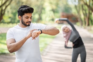 Handsome arabian guy in sport clothes standing at green park with smart watch on hand. Blur background of young woman in hijab warming up before training.