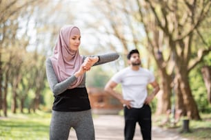 Young woman in hijab checking time on smart watch while muslim guy relaxing after training on background. Healthy sport couple exercising together at park.
