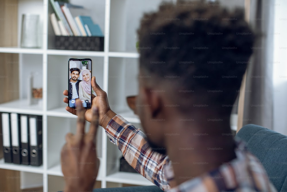 Afro american guy using modern smartphone for video call with happy muslim couple. Young man gesturing and talking while staying at home. Concept of isolation and remote communication.