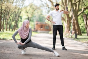 Fit young woman in hijab and sport outfit warming up at park before morning run. Handsome muslim guy standing on background and checking pulse on smart watch.