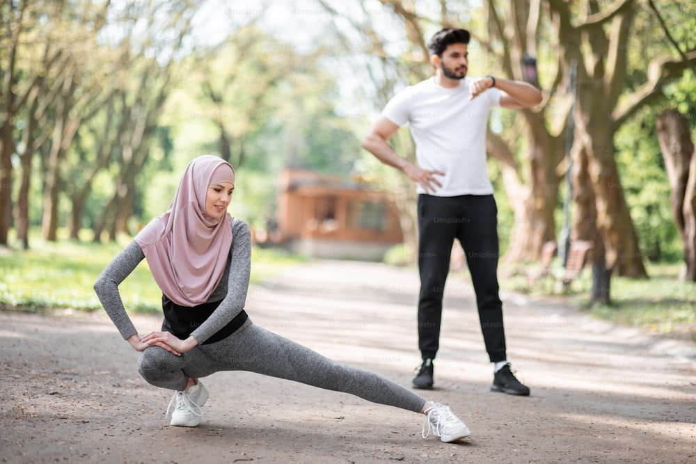 Mujer joven en forma con hiyab y atuendo deportivo calentando en el parque antes de correr por la mañana. Guapo musulmán de pie en el fondo y comprobando el pulso en el reloj inteligente.