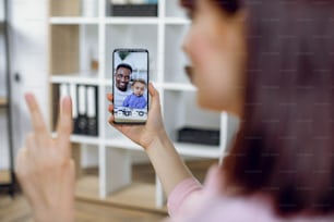 Caucasian woman having video call with african man that holding baby boy on hands. Young lady using modern smartphone for online communication while staying at home.