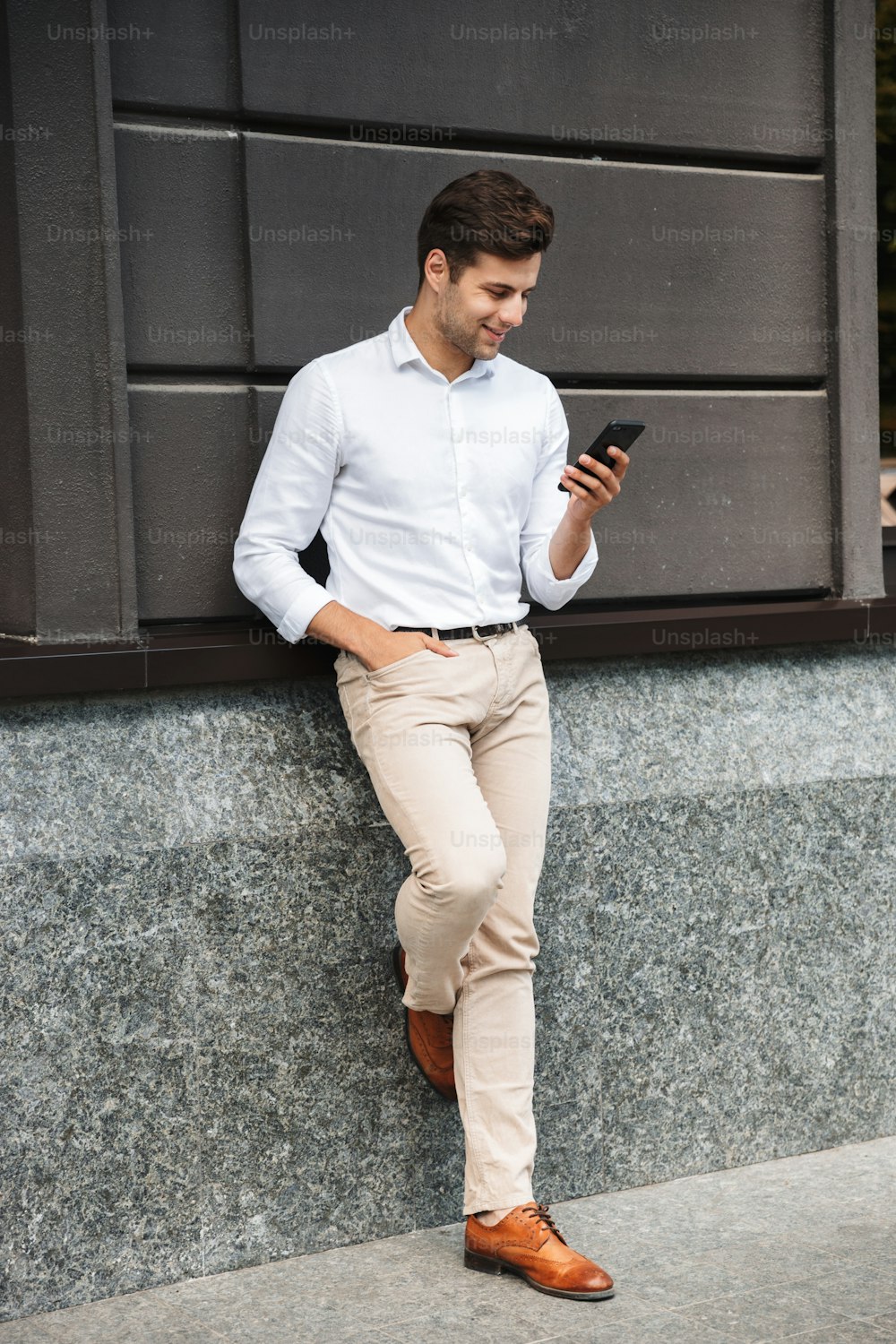 Attractive young male manager formal dressed leaning on a wall outdoors, using mobile phone