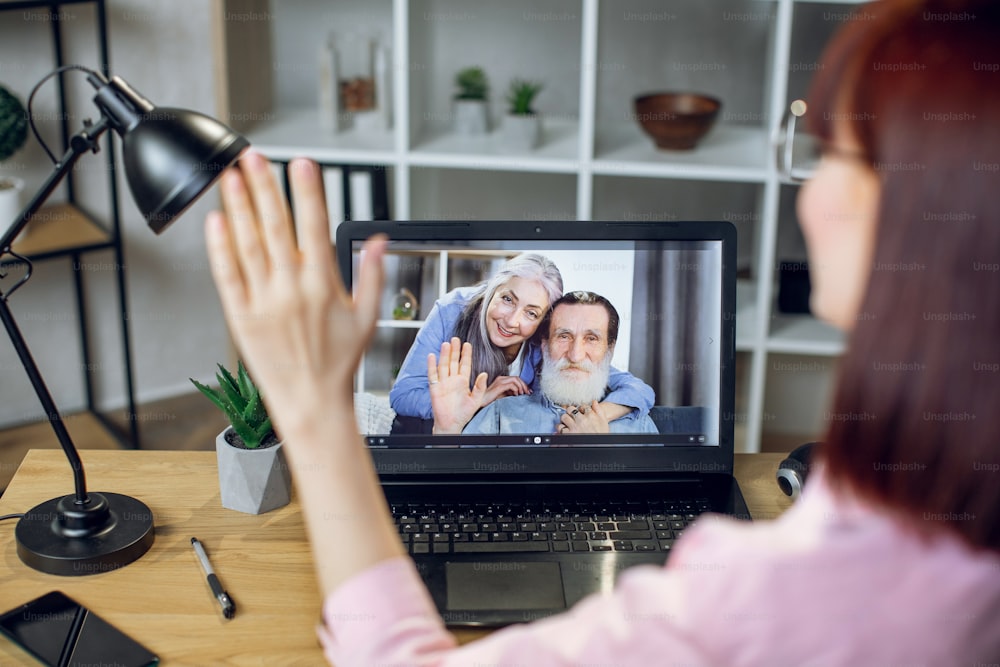 Pretty woman waving with hand during video conversation with grandparents. Young lady sitting at home and using modern laptop for distance communication.