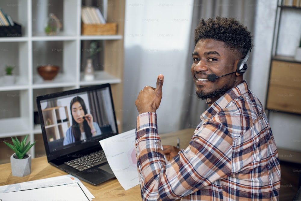 African man smiling and showing thumb up on camera while having video chat on laptop with asian woman. Two coworkers leading briefing remotely from home.