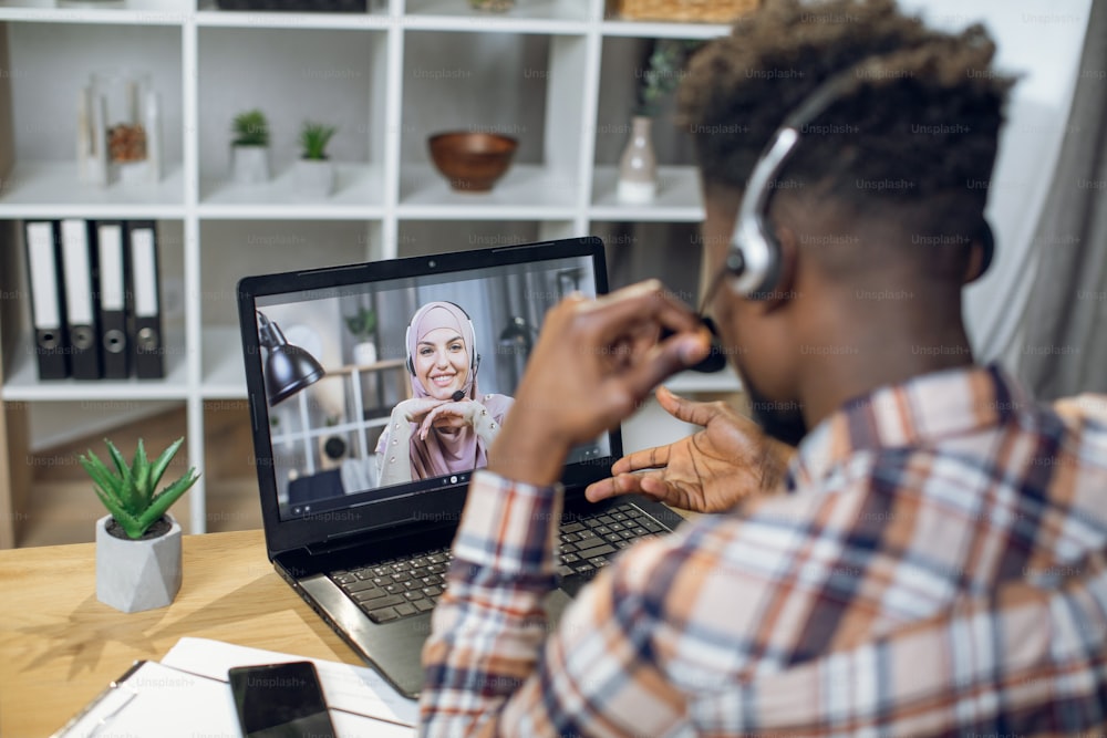 Vista de trás do cara afro-americano e da mulher muçulmana falando durante a chamada de vídeo enquanto fica em casa. Adorável casal usando fone de ouvido e laptop moderno para comunicação à distância.