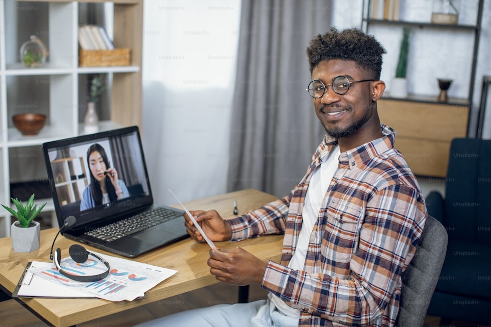 Hombre afroamericano feliz sentado a la mesa con una computadora portátil moderna y hablando con una mujer asiática a través de una videollamada. Concepto de trabajo remoto, tecnología y cooperación.