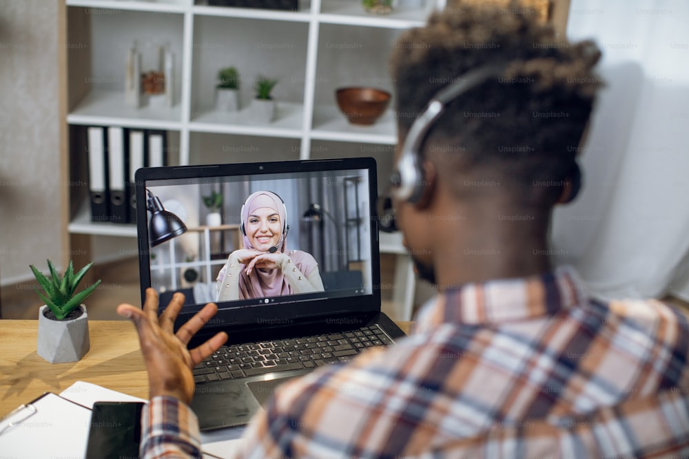 African man in headset having video chat on laptop with smiling muslim woman in hijab. Young couple communicating on distance during pandemic time.