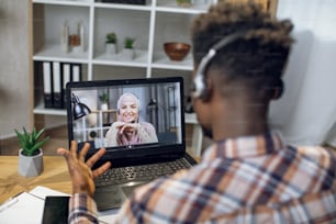 Hombre africano con auriculares que tiene videochat en una computadora portátil con una mujer musulmana sonriente con hiyab. Pareja joven que se comunica a distancia durante la pandemia.