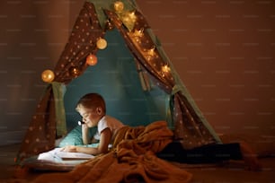 Lendo livro e usando lanterna. Menino em roupas casuais deitado perto da tenda à noite.