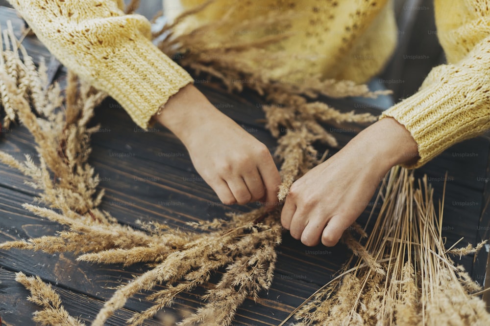 Florist in yellow sweater making rustic autumn wreath on dark wooden table. Hands holding dry grass and making stylish boho wreath with wildflowers and herbs on rustic wooden background.