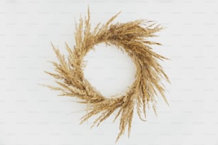 Stylish rustic boho wreath with dry grass hanging on white wall background. Boho autumn wreath with pampas grass isolated. Holiday workshop and fall decor