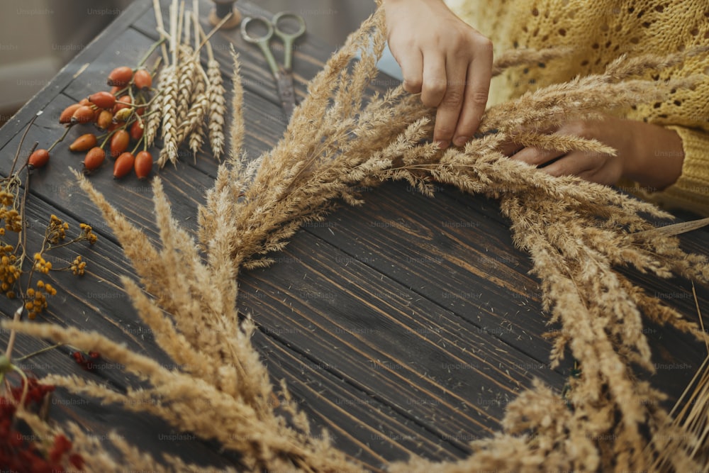 Making rustic autumn boho wreath. Hands holding dry grass and making stylish wreath with wildflowers, herbs and berries on rustic wooden background. Holiday workshop