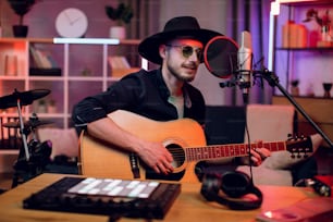 Handsome man singing in microphone and playing guitar while recording song at modern studio. Young singer in trendy hat and sunglasses creating new track indoors.