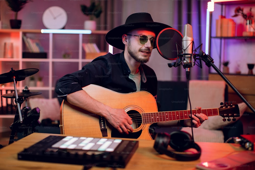 Bel homme chantant dans un micro et jouant de la guitare tout en enregistrant une chanson dans un studio moderne. Jeune chanteur portant un chapeau tendance et des lunettes de soleil créant un nouveau morceau à l’intérieur.