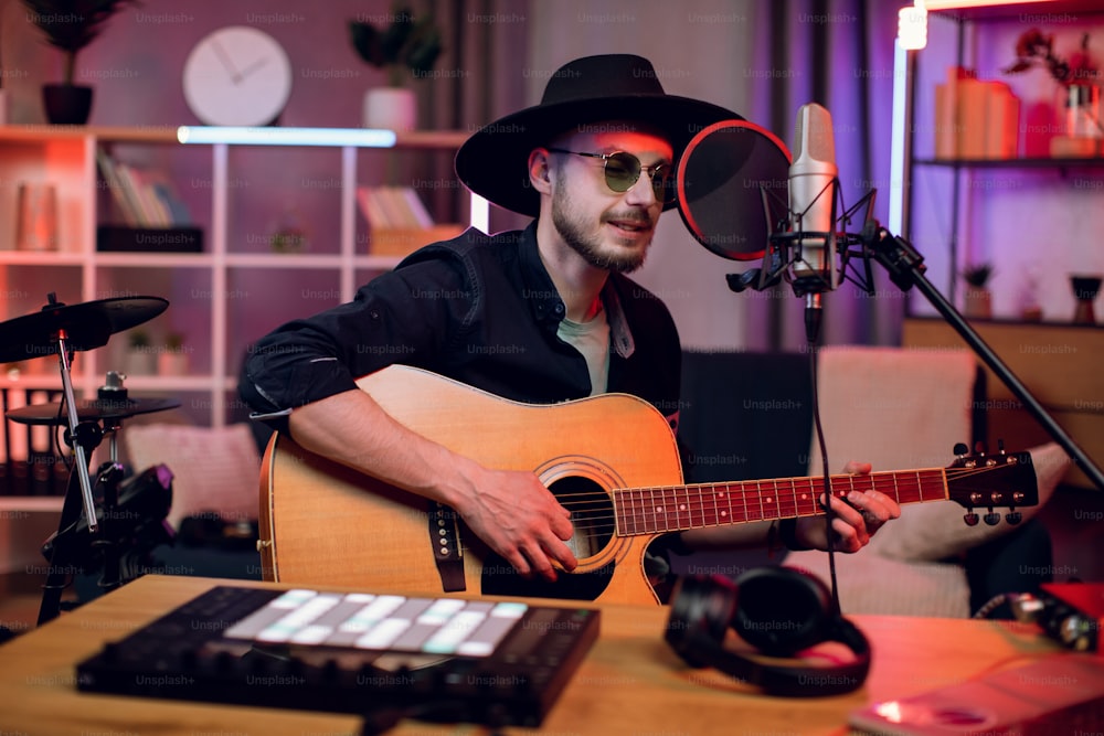 Hübscher Mann, der im Mikrofon singt und Gitarre spielt, während er im modernen Studio Songs aufnimmt. Junge Sängerin mit trendigem Hut und Sonnenbrille kreiert neuen Track drinnen.