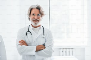Portrait of senior male doctor with grey hair and beard in white coat standing indoors in clinic.