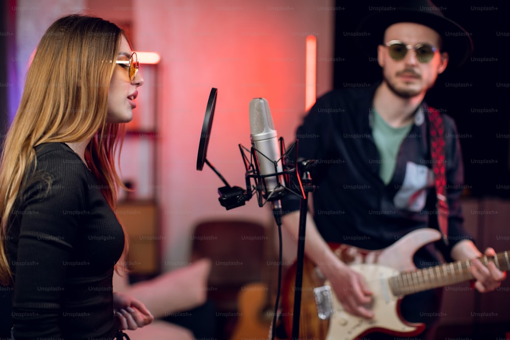 Young woman singing in microphone while man playing guitar near. Music band in stylish clothes creating hit music at modern studio. Art concept.