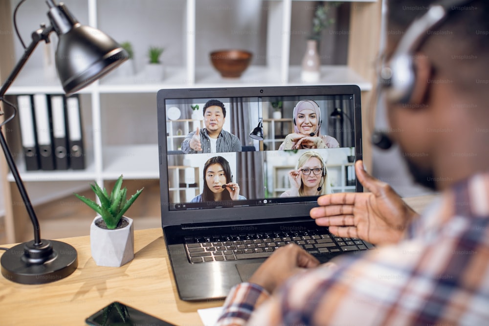Grupo multiétnico de personas que se reúnen de negocios a través de una videollamada en una computadora portátil. Vista desde el hombro de un hombre negro sentado en la oficina en casa y trabajando de forma remota.