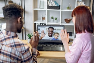 Young multicultural couple having video call on modern laptop with african man that holding cute baby boy on hands. Happy friends talking online while staying at home.