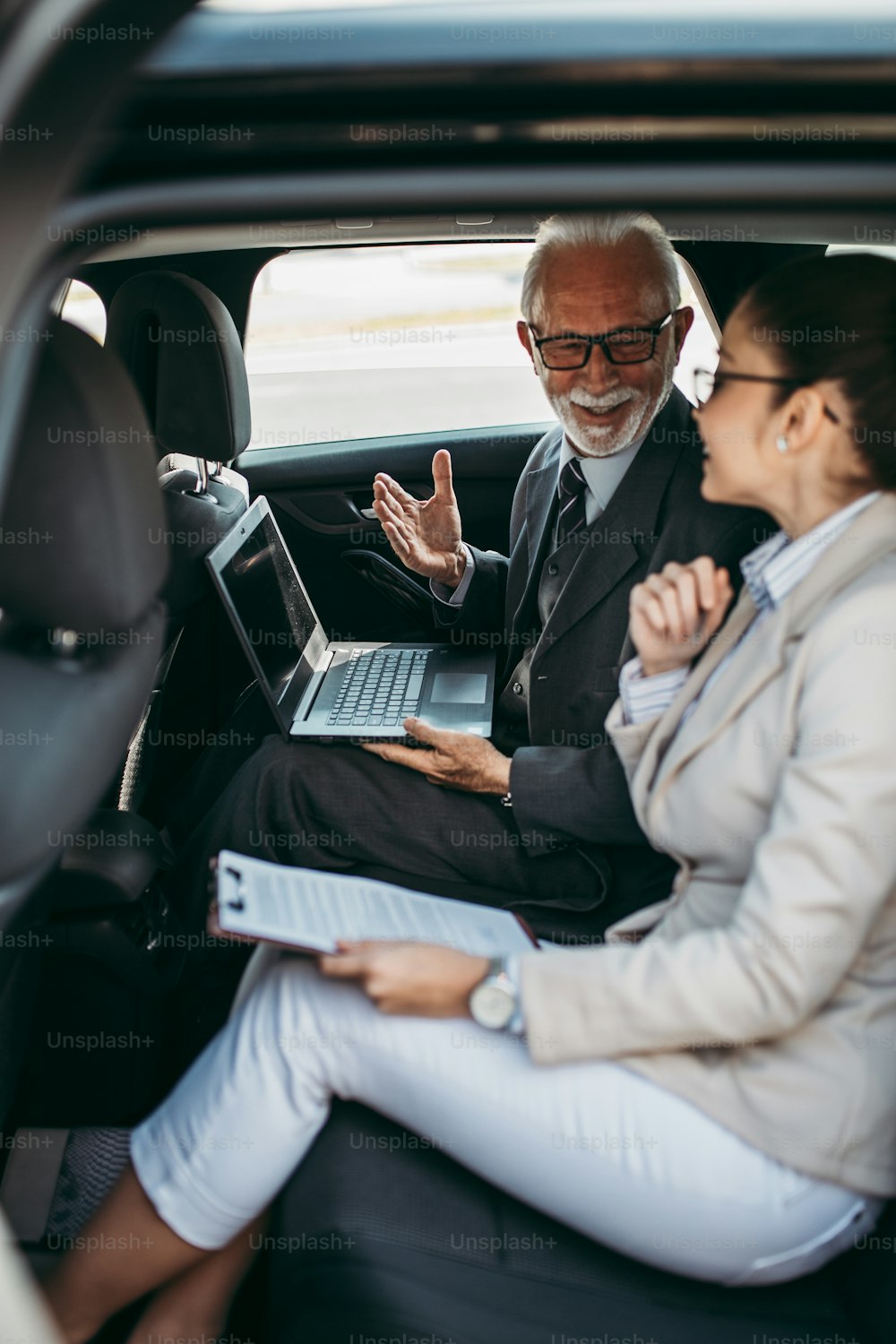 Un hombre de negocios de alto nivel y su colega o compañera de trabajo joven sentados en el asiento trasero de un automóvil de lujo. Hablan, sonríen y usan computadoras portátiles y teléfonos inteligentes. El transporte en el concepto de negocio corporativo.