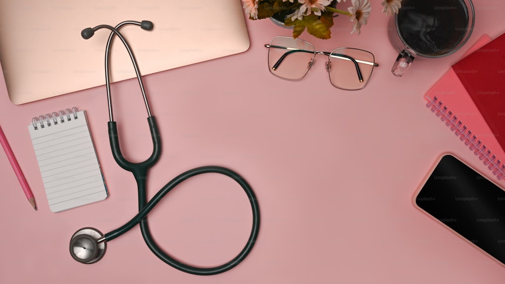 Stethoscope, smart phone computer laptop and glasses on pink background. Doctor workspace.
