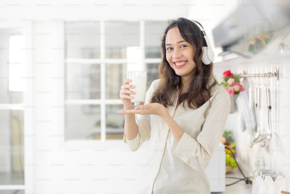 In a light modern kitchen, a beautiful woman drinks milk while listening to music. Before breakfast, she's having a good time and smiling.