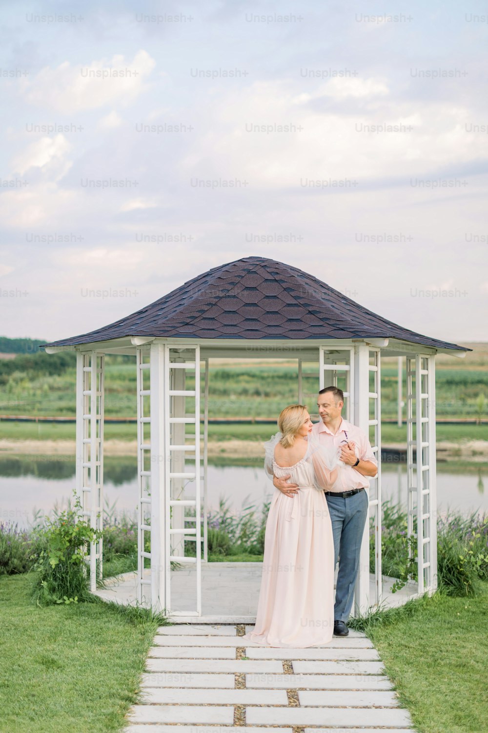 Romantic date on the shore of the pond. Happy mature couple, man and woman in elegant clothes, standing near wooden gazebo on the shore of beautiful lake
