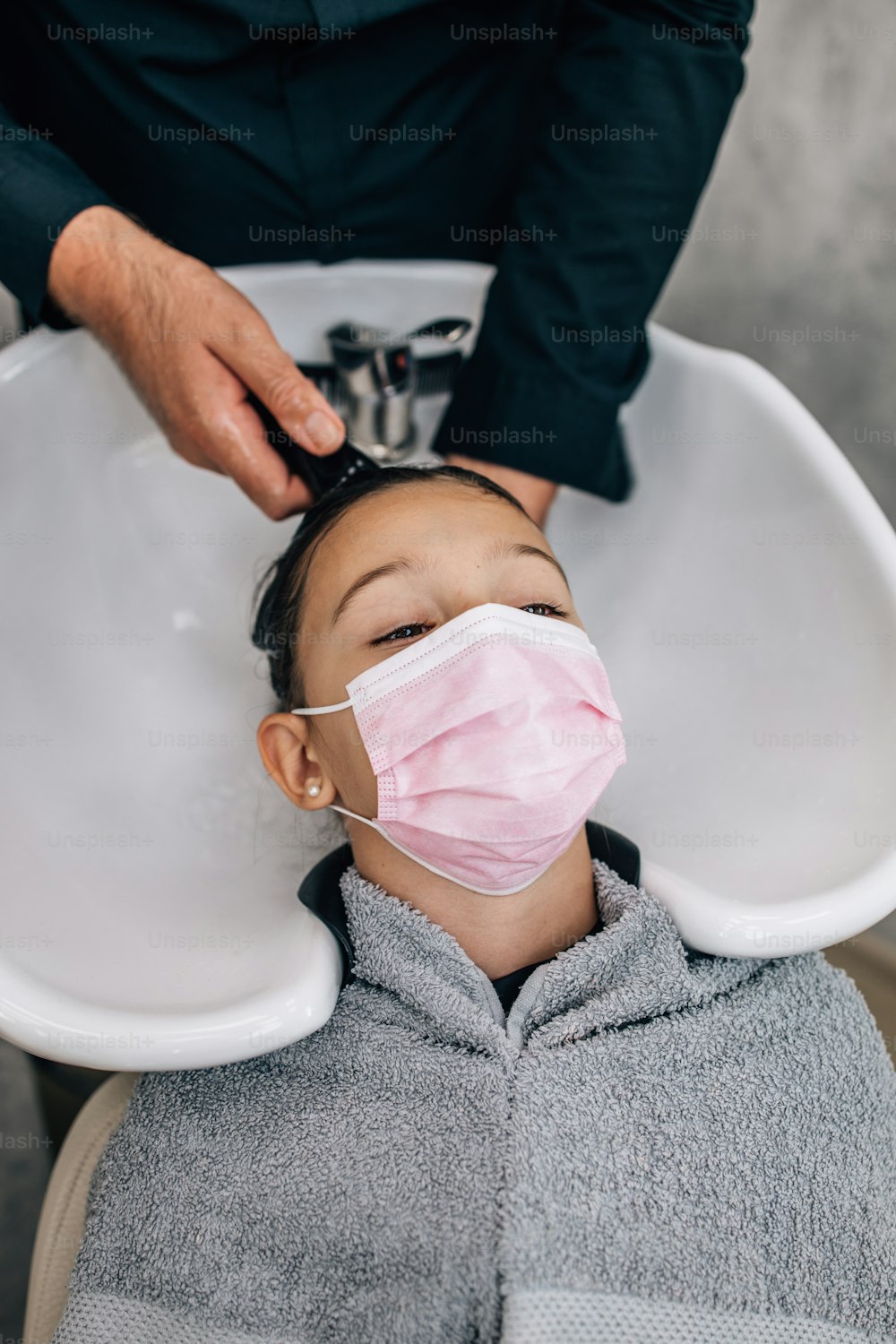 プロの美容師が優しく髪を洗っている間、髪型治療で保護フェイスマスクをした若い女の子。コロナウイルスのライフスタイルとニューノーマルのコンセプト。
