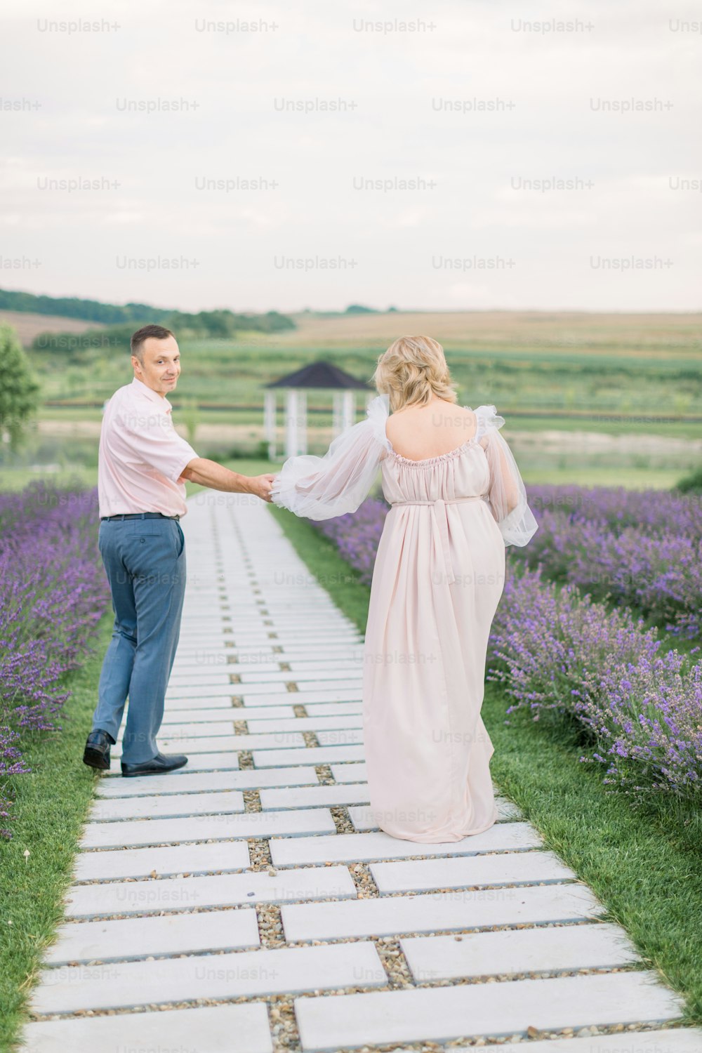 Outdoor shot of happy romantic mature couple in love walking on pathway through flowering lavender field, holding hands and enjoying moments together