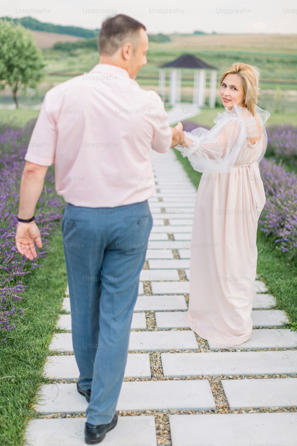 Give me your hand, follow me. Love through the years together. Pretty charming blond mature lady in elegant dress, holding hand and walking with her handsome man outdoors in lavender field with gazebo