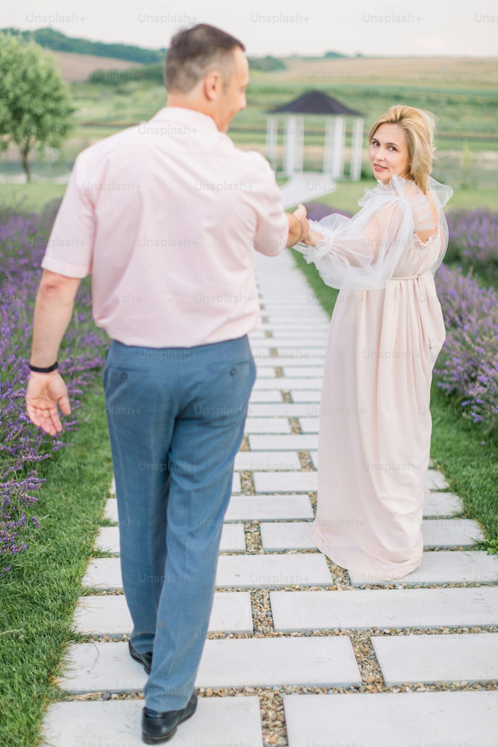 Donne-moi la main, suis-moi. L’amour à travers les années ensemble. Jolie charmante dame blonde mature en robe élégante, se tenant la main et marchant avec son bel homme à l’extérieur dans le champ de lavande avec gazebo