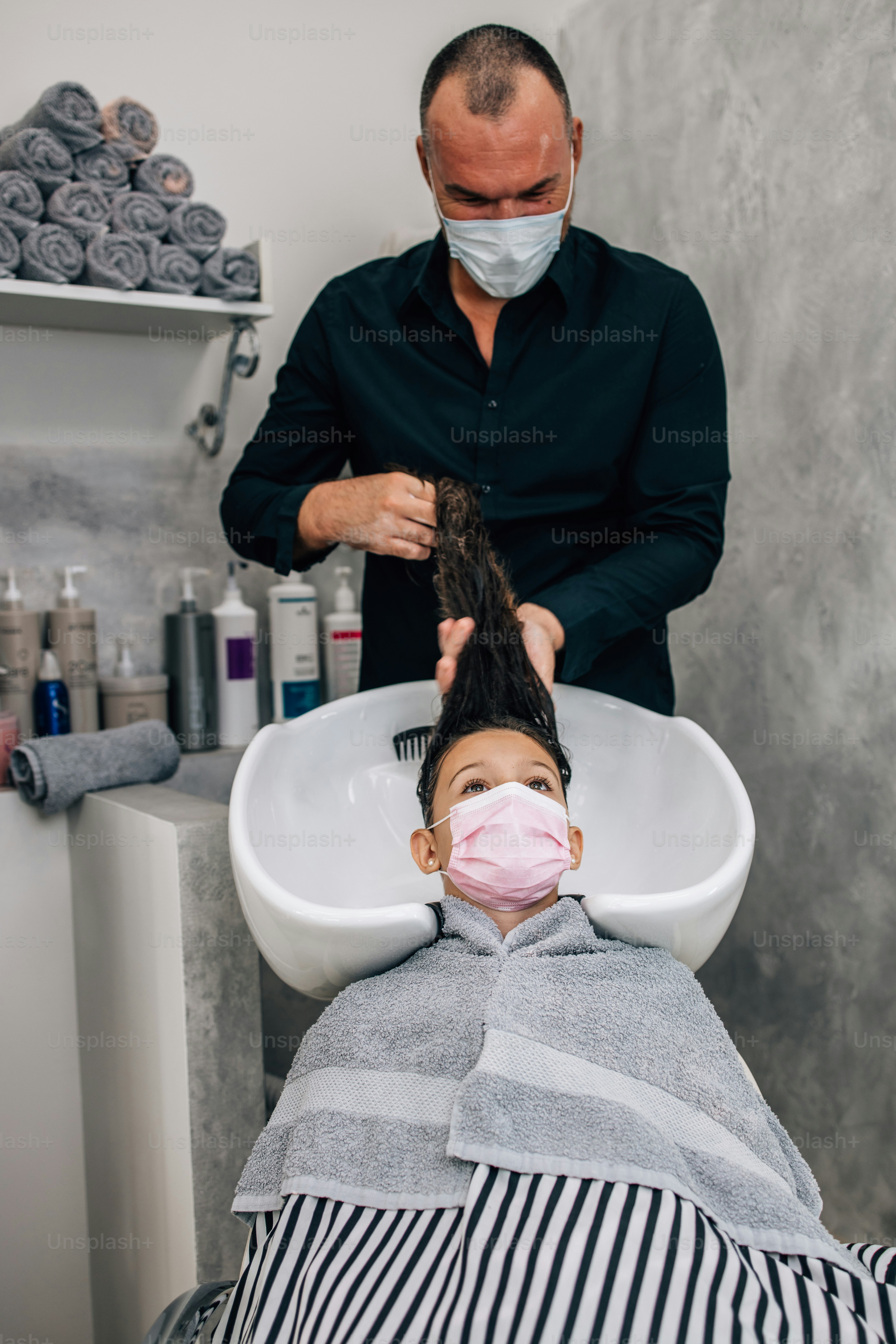Young girl with protective face mask at hairstyle treatment while professional hairdresser gently washing her hair. Coronavirus lifestyle and new normal concept.