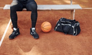 Sits with black bag and preparing for the game. African american man plays basketball on the court outdoors.