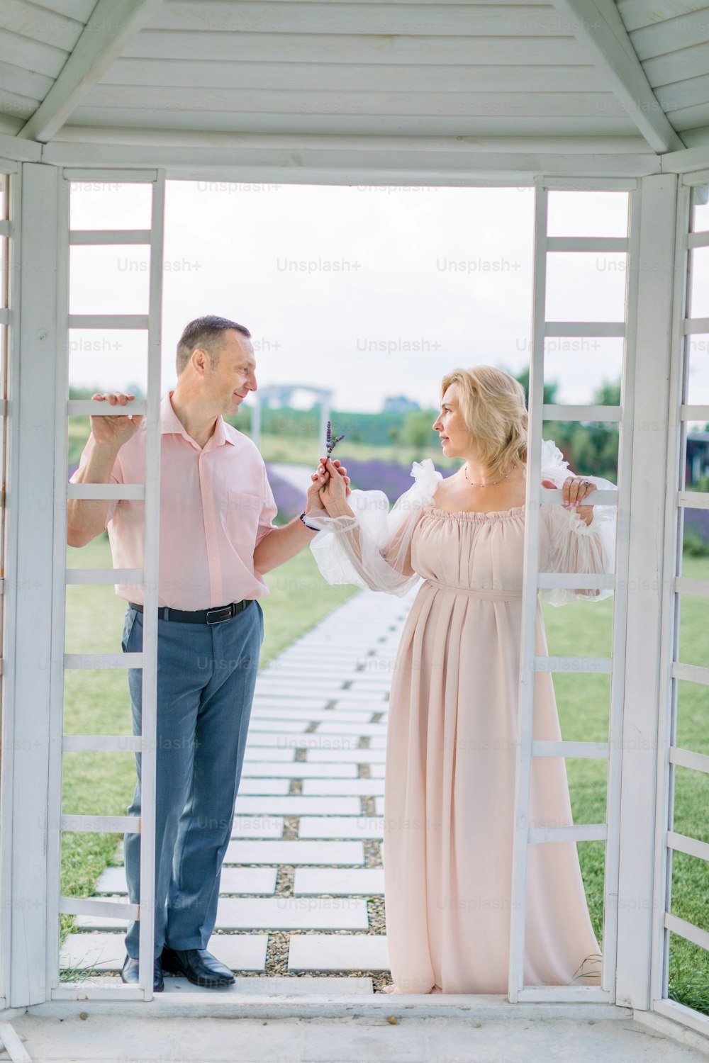 Ganzkörperporträt eines reifen, eleganten Paares, das außerhalb eines weißen Holzpavillons steht, sich gegenseitig anschaut und blühenden Lavendel in den Händen hält
