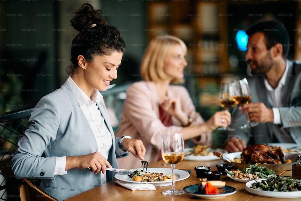 Young businesswoman enjoying while eating lunch in a restaurant. Her colleagues are toasting with wine in the background.