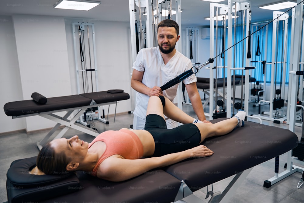 Young woman training leg muscles and joints under doctors supervision on massage table in rehabilitation center. Male physical therapist assists patient in recovery from sports injury. Rehab clinic