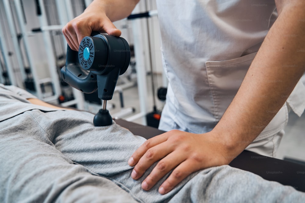 Faceless physical therapist with massage percussion gun treats girls leg close-up. Muscle stimulator treatment of relieved leg muscles in rehabilitation clinic