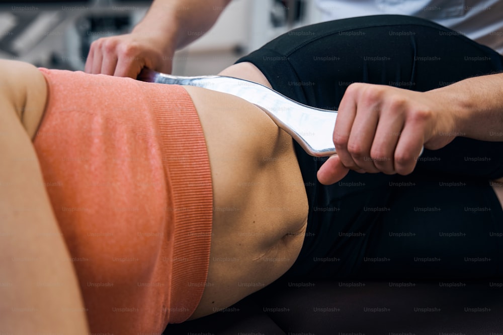 물리 치료를 위해 옆으로 누워 있는 여성의 클로즈업. 척추 지압사는 IASTM 도구로 복근을 누릅니다. 스포츠 부상을위한 치료 마사지. 연조직 치료제. 대체 의학