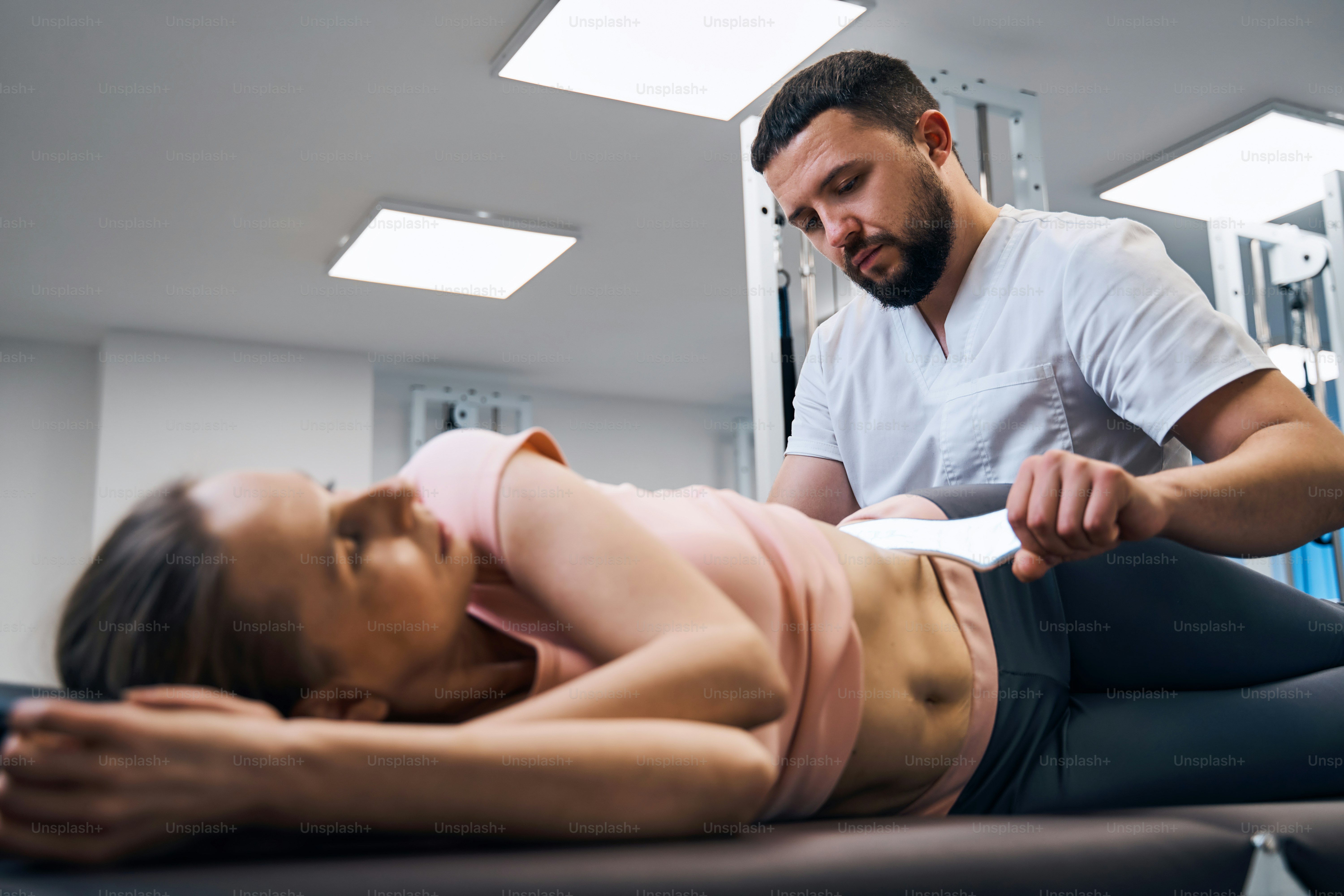 Woman lying on treatment bed physiotherapist presses her abs with IASTM instrument side view. Therapeutic massage for sports injury. Remedial therapy pressure using soft tissue instrument. Rehab