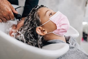 Junges Mädchen mit schützender Gesichtsmaske bei der Frisurenbehandlung, während professionelle Friseurin sanft ihre Haare wäscht. Coronavirus-Lebensstil und neues normales Konzept.