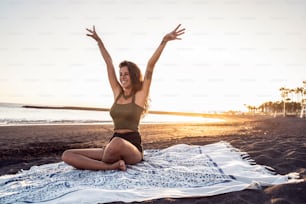 Happy carefree beautiful woman enjoying sunset evening on the sandy beach, having fun, smiling, sitting on the blanket. Summer positive vibes. Real people emotions, lifestyle.
